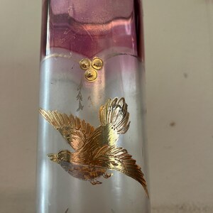May include: A tall, clear glass vase with a gold rim and a frosted silver body. The top section is a deep purple. A gold bird in flight is depicted on the vase, along with gold floral accents. The vase is likely for decorative purposes.