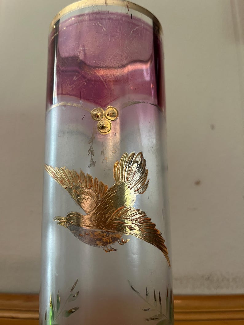May include: A tall, clear glass vase with a gold rim and a frosted silver body. The top section is a deep purple. A gold bird in flight is depicted on the vase, along with gold floral accents. The vase is likely for decorative purposes.