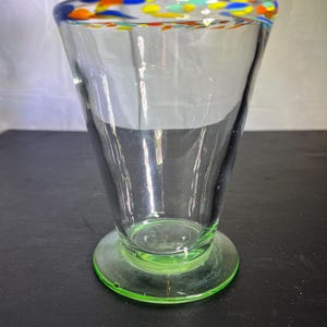 May include: A clear glass vase with a flared shape and a wide, round base. The rim is decorated with colorful specks of blue, orange, yellow, and green. The base is a bright green color.