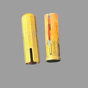 May include: Two vintage brass pen holders. One is a solid, fluted cylinder with a vertical slot. The other is a hollow cylinder with a vertical slit, revealing an orange interior. Both have a worn, aged appearance.