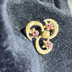 May include: A gold-toned brooch with a three-part design. Each section features a crescent shape adorned with small, round pearls. The brooch incorporates three red gemstones and a central pearl. The design includes star accents.