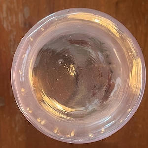 May include: A clear glass with a slightly pinkish hue, viewed from above. The rim of the glass has a subtle curve, and the interior appears to contain a light-colored liquid. Small flecks of gold are visible inside the glass.