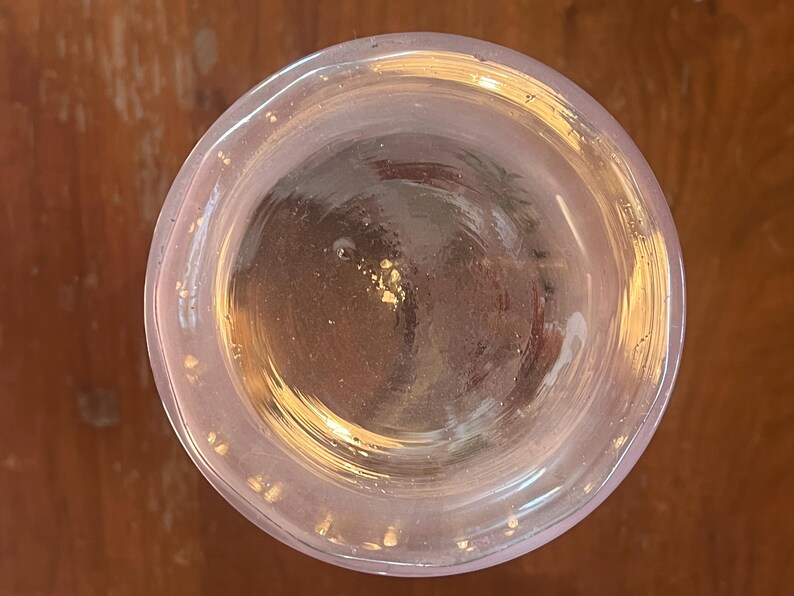 May include: A clear glass with a slightly pinkish hue, viewed from above. The rim of the glass has a subtle curve, and the interior appears to contain a light-colored liquid. Small flecks of gold are visible inside the glass.