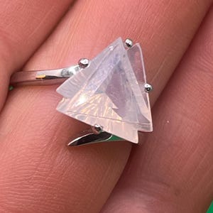 May include: A silver-toned ring featuring a large, triangular, translucent pale pink stone with a faceted surface. The stone is secured by three small metal prongs on a polished metallic band that wraps around the finger.