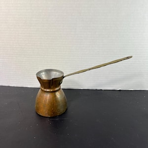 May include: A small, copper-colored Turkish coffee pot with a long, thin handle. The pot has a rounded base and a narrow neck, with a silver-colored interior. The handle is made of a lighter-colored metal.