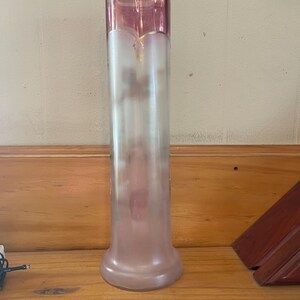 May include: A tall, elegant glass vase with a flared base and a cylindrical body. The top section is a deep pink, transitioning to a frosted, translucent body. A gold rim accents the top. The vase is approximately 12 inches tall.