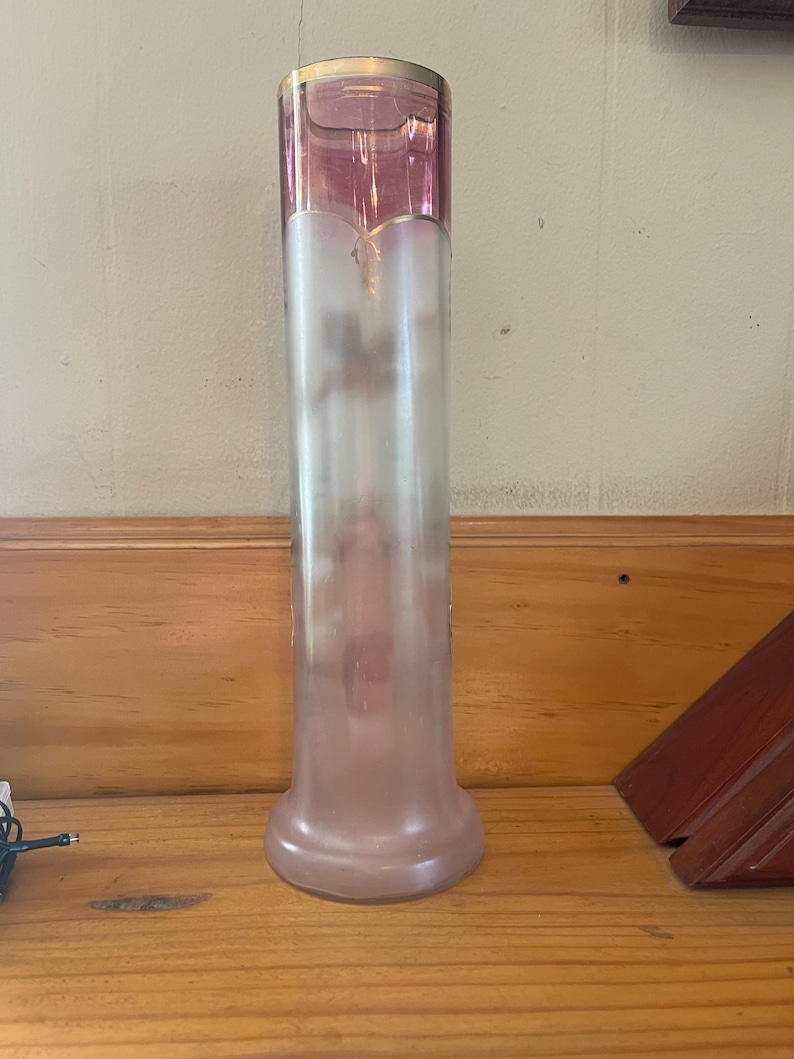 May include: A tall, elegant glass vase with a flared base and a cylindrical body. The top section is a deep pink, transitioning to a frosted, translucent body. A gold rim accents the top. The vase is approximately 12 inches tall.