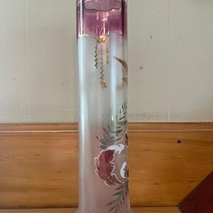 May include: Tall, cylindrical glass vase with a flared base and a gold rim. The top section is a deep pink, transitioning to a frosted white body. Features hand-painted floral and leaf designs in red, green, and gold. A decorative piece.