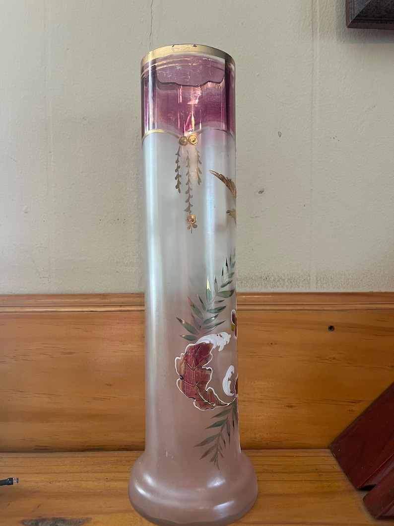 May include: Tall, cylindrical glass vase with a flared base and a gold rim. The top section is a deep pink, transitioning to a frosted white body. Features hand-painted floral and leaf designs in red, green, and gold. A decorative piece.