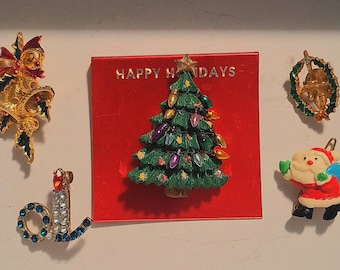 May include: A collection of Christmas-themed brooches. Includes a gold bell with red bow, a Christmas tree on a red card with "HAPPY HOLIDAYS" text, a wreath, a Santa Claus, and a jeweled candle.
