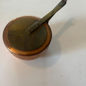 May include: A small copper saucepan with a brass handle. The pan has a rounded base and a flat, circular top. The handle is attached with rivets and has a curved end. The pan is likely vintage and shows signs of wear.