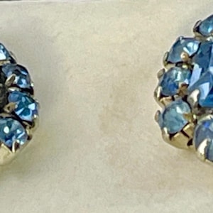 May include: A pair of vintage earrings featuring a central, square-cut, light blue gemstone surrounded by smaller, round, light blue stones. The earrings have a gold-colored metal setting and a classic, elegant design.