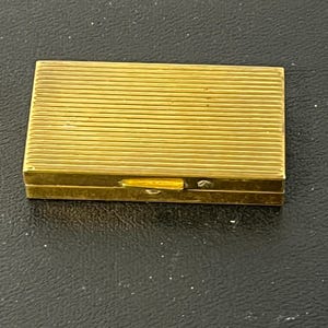 May include: A rectangular, gold-colored pillbox with a ribbed design on the top and a small clasp. The box is closed and appears to be made of metal. The pillbox is on a dark surface.