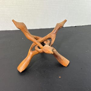 May include: A handcrafted wooden stand with a unique, interwoven design. The stand is made of light brown wood and features four curved legs that intersect in the center. It is designed to hold a small object.