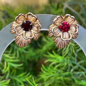 May include: A pair of gold-toned metal earrings shaped like flowers with textured petals and a single round, deep red faceted stone set in the center of each bloom. The design features layered petals and leaves with a metallic finish.