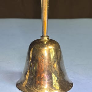 May include: A shiny, gold-colored bell with a long, slender handle. The bell has a rounded base and a smooth, polished surface. The handle is cylindrical with horizontal grooves near the top. The bell is likely made of metal and reflects light.