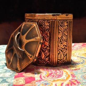 May include: A vintage copper-colored metal box with an ornate floral design. The box has a hinged lid with a decorative handle. The box is sitting on a colorful patterned fabric.