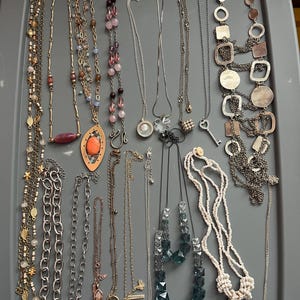 May include: A collection of necklaces in various styles and lengths, displayed on a gray surface. The necklaces feature a variety of materials, including beads, chains, and pendants. Colors include silver, gold, and various shades of blue, pink, and brown. Some necklaces have unique charms.