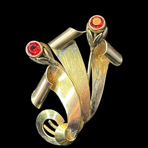 May include: A vintage gold-toned brooch with a swirling, ribbon-like design. The brooch features three red gemstone accents, adding a pop of color against the metallic backdrop. The design is abstract and sculptural.