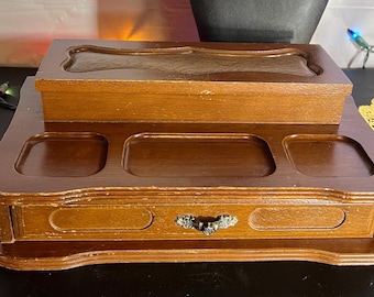May include: A vintage wooden jewelry box with a dark brown finish. The box has a tiered design with a top lid, three recessed compartments, and a drawer with a decorative metal handle. The box is designed to store jewelry.