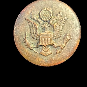 May include: A vintage, circular button with a detailed embossed design. The button features a central emblem of an eagle with outstretched wings, a shield, and a wreath. The button has a bronze color and a slightly aged appearance.