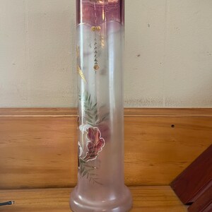 May include: A tall, cylindrical vase with a frosted glass body and a flared base. The top section is a deep pink color with a gold rim. The vase features painted floral and leaf designs in shades of red, green, and gold.