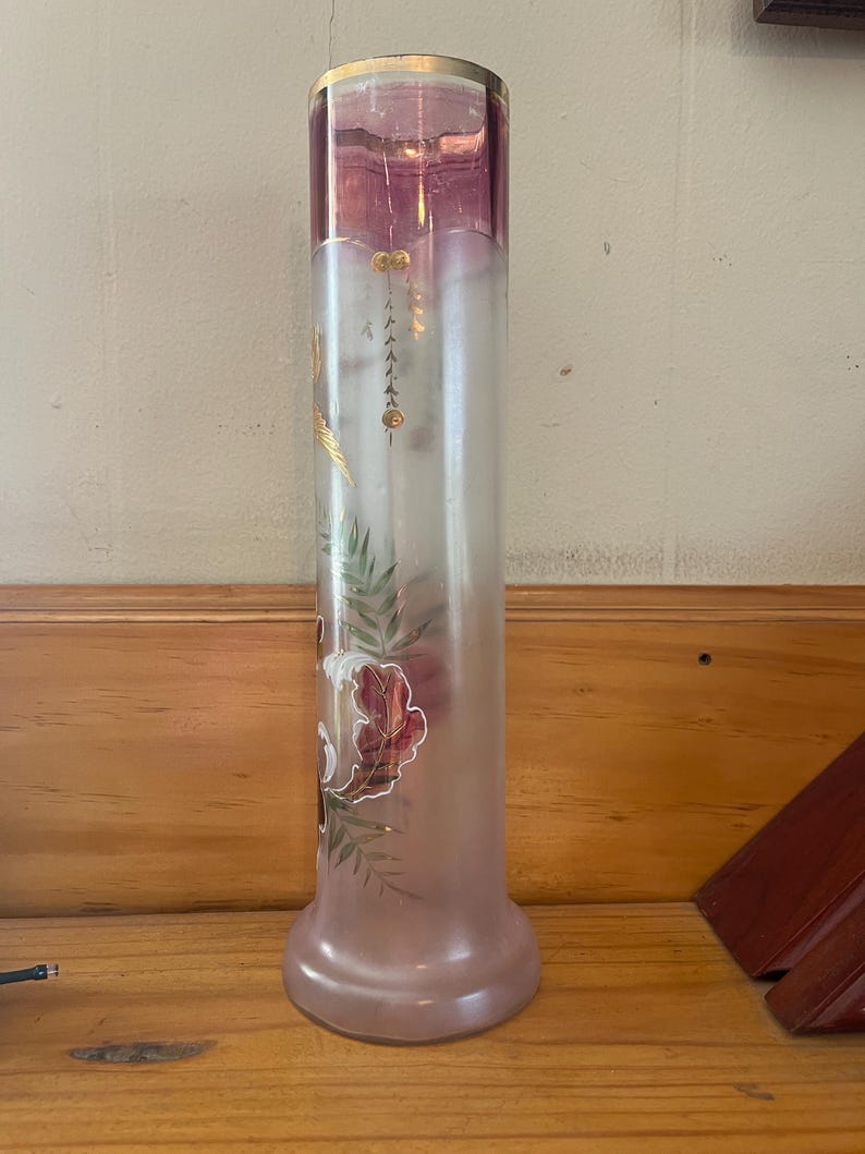 May include: A tall, cylindrical vase with a frosted glass body and a flared base. The top section is a deep pink color with a gold rim. The vase features painted floral and leaf designs in shades of red, green, and gold.