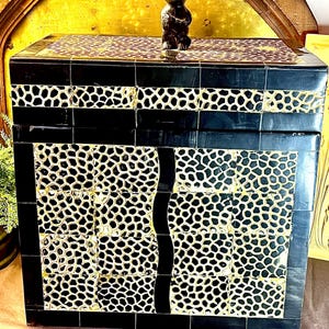 May include: A rectangular box with a hinged lid, featuring a black and white giraffe-like animal print pattern. The box is accented with solid black borders and a vertical wavy black stripe. A small, dark brown carved monkey figurine sits on top.
