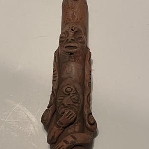 Puede incluir: Un tótem de madera tallada de color marrón rojizo con dos caras y un agujero en la parte superior. Las caras tienen rasgos detallados, y la cara inferior tiene brazos envueltos alrededor. El tótem es probablemente un objeto decorativo o ceremonial.