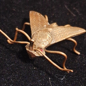 May include: A bronze insect figurine with intricate details, featuring a detailed wing design and a red gemstone accent. The insect is posed with its legs outstretched.