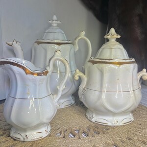 May include: A white porcelain tea set with gold trim. The set includes a teapot, sugar bowl, and creamer. The teapot has a lid and a curved handle. The sugar bowl has a lid and two handles. The creamer has a handle and a spout.