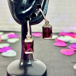 May include: A pair of silver drop earrings with rectangular, deep red gemstones. The earrings are displayed on a black stand, with a blurred background of pink and white heart-shaped confetti.