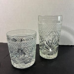 May include: Two clear, cut-glass drinking glasses. One is a short, round tumbler, and the other is a taller, cylindrical glass. Both feature a diamond and leaf pattern. The glasses are empty and sit on a dark surface.