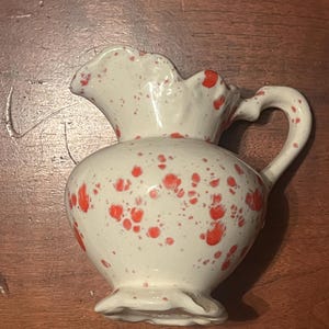 May include: A small, white ceramic pitcher with a ruffled rim and handle. The pitcher is decorated with red speckles. The base of the pitcher has a scalloped edge. The pitcher is sitting on a wooden surface.