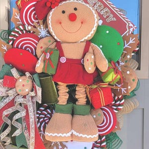 May include: A festive Christmas wreath featuring a plush gingerbread person with a red dress and green boots. The wreath is adorned with candy, gingerbread cookies, and a sign that says "MERRY".