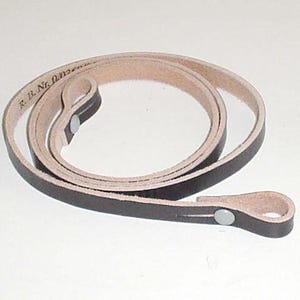Op de afbeelding: Een opgerolde leren riem met een donkerbruine buitenkant en een lichtere binnenkant. De riem heeft aan één uiteinde een lus en is vastgezet met een metalen klinknagel. De tekst "R. B. Nr. 0" is zichtbaar.