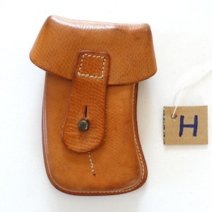 Könnte beinhalten: Eine hellbraune Ledertasche mit geschwungener Form, die über eine Umschlagklappe mit zentralem Metallknopf und hellen Nähten verfügt. Ein kleines quadratisches Pappschild mit dem handgeschriebenen Buchstaben H in blauer Tinte hängt an einer weißen Schnur.