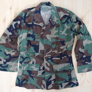 Könnte beinhalten: Eine langärmelige Tarnjacke mit Waldmuster in den Farben Dunkelgrün, Braun, Schwarz und Hellgrün. Die Jacke verfügt über einen spitzen Kragen, eine Knopfleiste auf der Vorderseite und vier rechteckige Vordertaschen mit geknöpften Klappen.