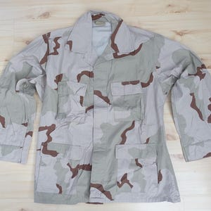 May include: A long-sleeved button-up jacket featuring a desert camouflage pattern in shades of tan, light brown, and dark brown. The jacket includes a pointed collar, four front pockets with buttoned flaps, and adjustable buttoned cuffs on sleeves.