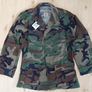 Könnte beinhalten: Eine langärmelige Camouflage-Jacke mit Waldmuster in Grün, Braun und Schwarz. Die Jacke verfügt über einen Kragen, geknöpfte Manschetten und vier Vordertaschen mit Klappen. Der Text PARKS und U.S. ARMY ist auf der Brust eingestickt.