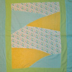 May include: A green, yellow, and blue patchwork quilt with a white background. The quilt has a polka dot pattern, a gingham pattern, and a solid yellow pattern. The quilt is framed with a green border.