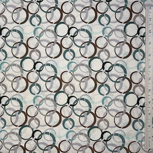 May include: Fabric with a repeating pattern of overlapping circles in shades of brown, gray, teal, and black on a cream background. The circles vary in size and are arranged in a random, abstract design. A ruler is visible on the right side.