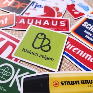 Könnte beinhalten: Eine Sammlung verschiedener rechteckiger und quadratischer Aufkleber mit diversen Markenlogos, grafischen Designs und deutschen Texten wie AUHAUS, SCHMERZ und STABIL BRUDER. Die Aufkleber sind überlappend angeordnet und weisen leuchtende Farben auf.