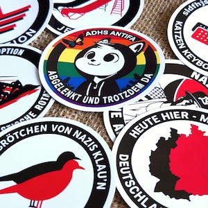 Może przedstawiać: Kolekcja okrągłych naklejek winylowych z różnymi wzorami graficznymi, w tym centralną naklejką z kotem z kreskówki, tęczowym tłem i napisem ADHS ANTIFA ABGELENKT UND TROTZDEM DA, oraz innymi naklejkami z symbolami politycznymi i tekstem.