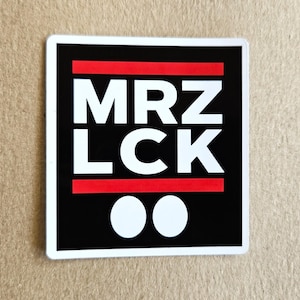 Op de afbeelding: Vierkante sticker met de witte tekst "MRZ LCK" op een zwarte achtergrond. Het heeft twee rode horizontale lijnen en twee witte cirkels. De sticker heeft een witte rand.