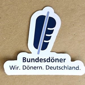 Könnte beinhalten: Ein weißer Aufkleber mit einer dunkelblauen Grafik eines vertikalen Döner-Spießes. Unter der Grafik steht der Text "Bundesdöner" in der ersten Zeile und "Wir. Dönern. Deutschland." in der zweiten Zeile in dunkelblauer serifenloser Schrift.