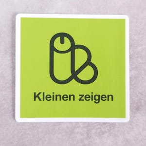 Peut inclure: Un autocollant carré avec un fond vert citron et une bordure blanche. Au centre, un graphique en ligne noire représente une forme phallique. Sous le graphique, le texte noir indique "Kleinen zeigen" dans une police sans empattement.