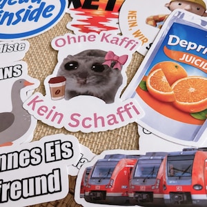 Op de afbeelding: Een verzameling diverse vinylstickers met verschillende ontwerpen, waaronder een hamster met een roze strik die een koffiebeker vasthoudt met de tekst Ohne Kaffi Kein Schaffi, een sapzakje met het label DepriSun Juicide, een gans en een rode trein.