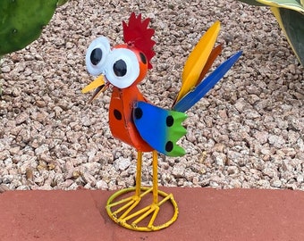 Può includere: Una statuetta di gallo in metallo colorato con grandi occhi bianchi, accenti rossi, arancioni, gialli, blu e verdi. Il gallo è in piedi su una base in metallo gialla.