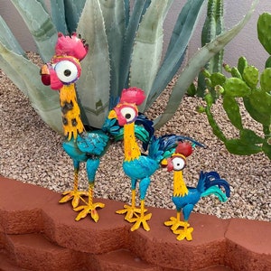 May include: Three colorful metal rooster figurines with blue, yellow, and red accents. The roosters are standing on a brown surface with green plants in the background.